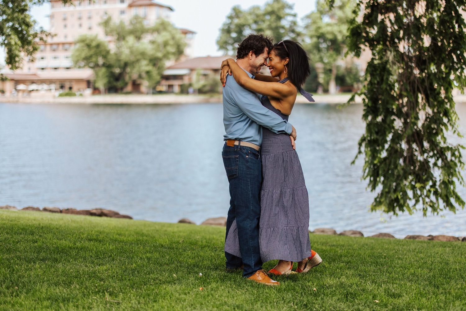 Couple sits together on grassy lakeside hill overlooking resort buildings on a peaceful summer afternoon.