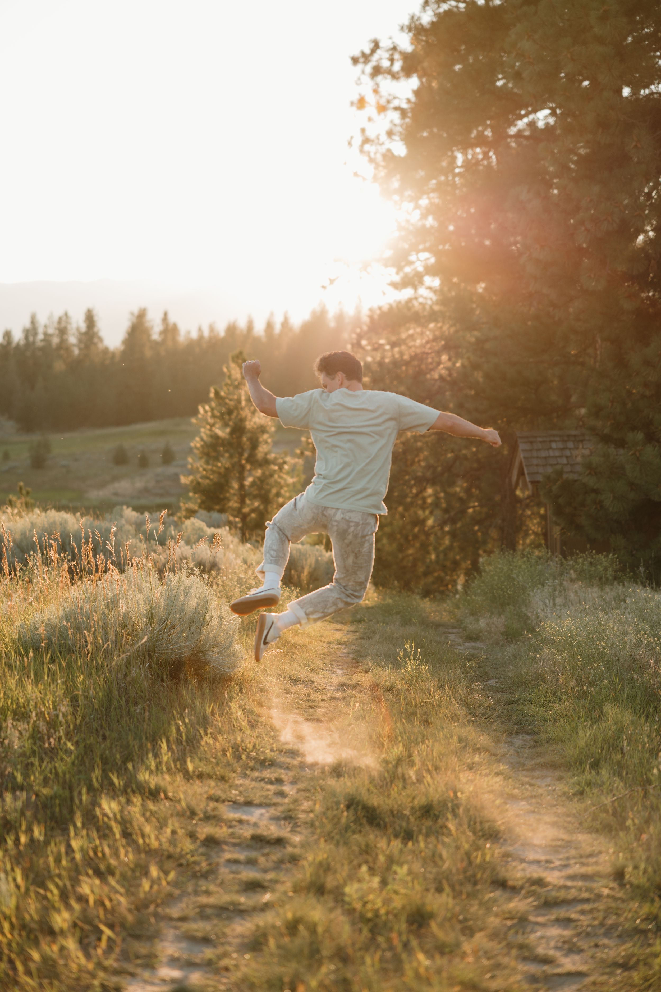 Person in white clothing jumps joyfully on a dirt trail during golden hour sunset with evergreen forest backdrop.