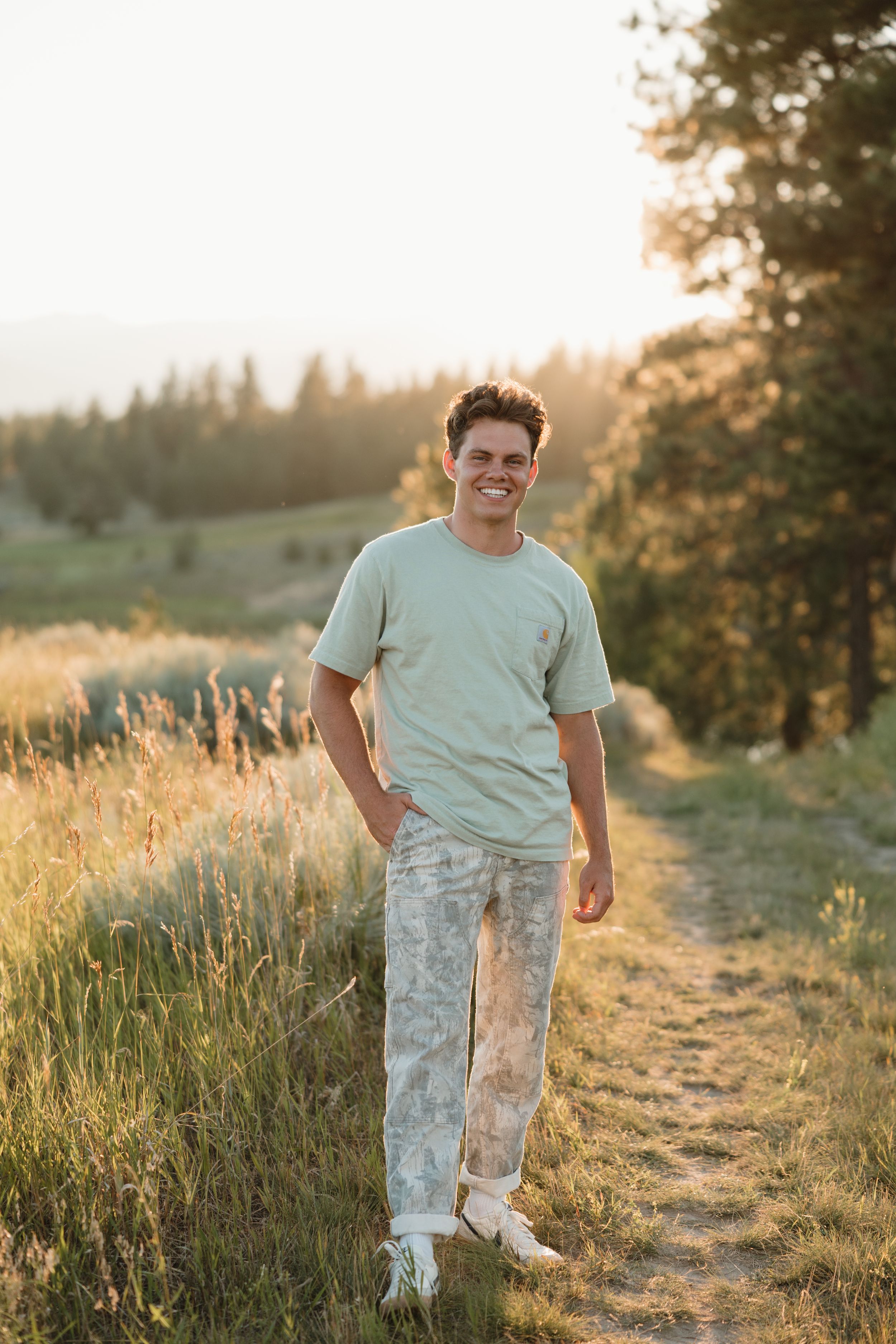 A person stands on a dirt trail at sunset wearing a mint green t-shirt and light-colored pants in a grassy field.