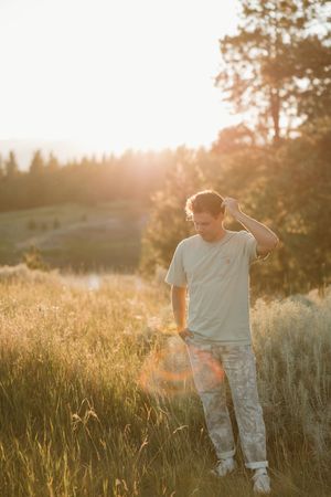 Person standing in golden field at sunset with mountains and trees in background against warm evening light.