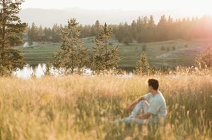 Sunlit figure sits in golden meadow beside tranquil lake at sunset with evergreen forest in background.