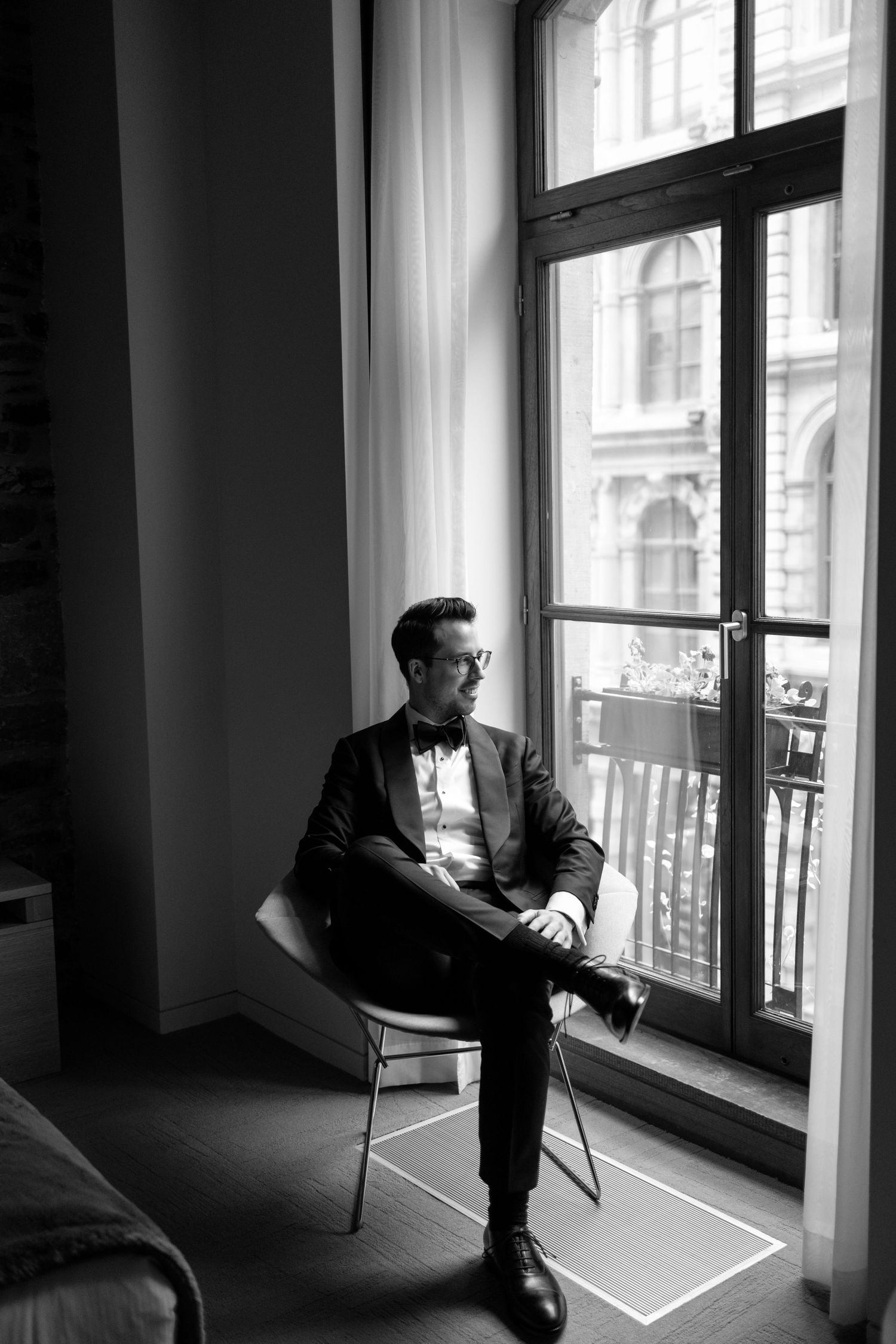 Editorial black and white photo of the groom in a tuxedo sitting by a large window captured by Montreal wedding photographer, Aliza Dworkind