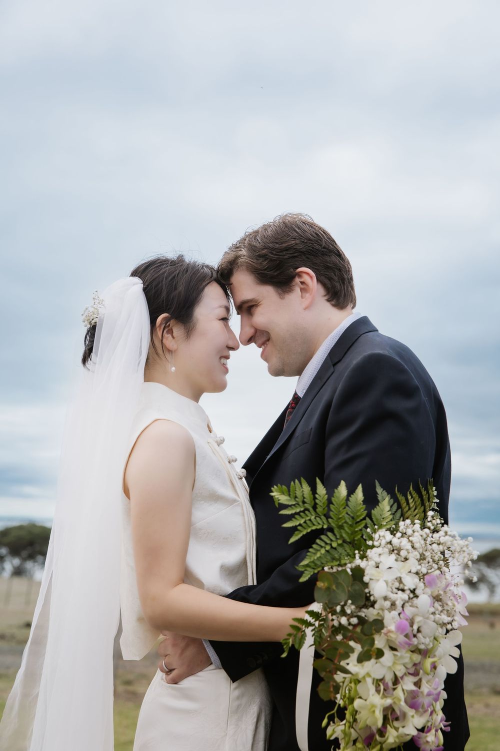A romantic wedding couple shares an intimate moment outdoors with a bridal bouquet of flowers against cloudy skies.