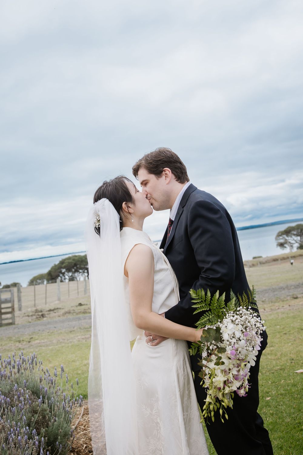 A romantic outdoor wedding portrait with the couple sharing a kiss near lavender flowers and a coastal backdrop.