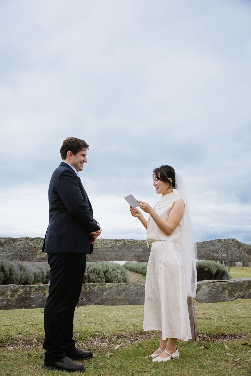 Newlyweds stand in a grassy field reading wedding vows to each other with mountains in the background.