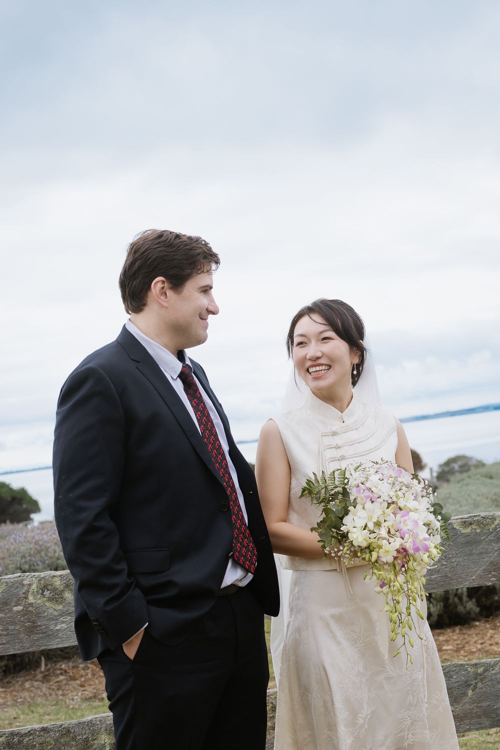 Newlyweds share a tender moment in front of a scenic coastal landscape while holding a white floral bouquet.