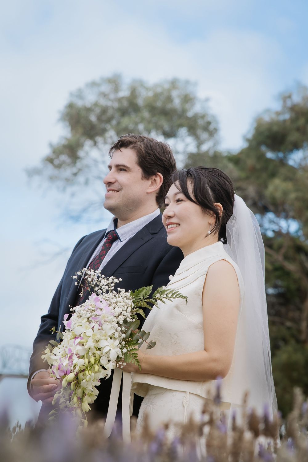 A happy couple poses outdoors on their wedding day with a beautiful floral bouquet against a cloudy sky.
