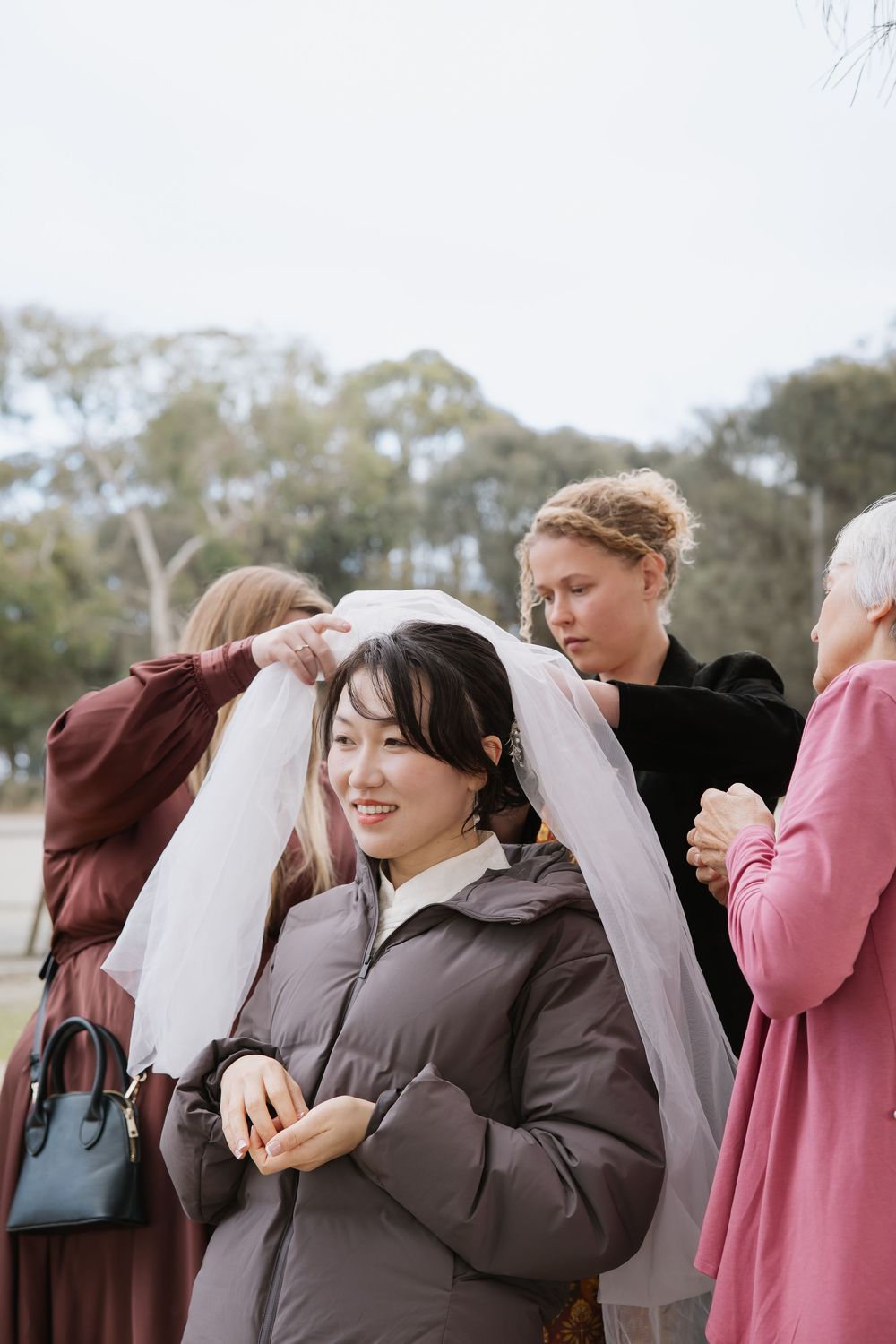 Helpers adjust a wedding veil on a smiling woman wearing a brown winter coat outdoors.