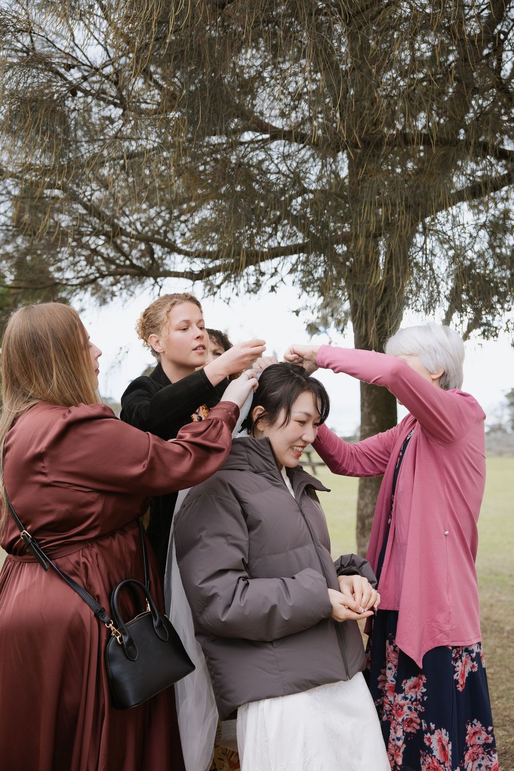 Wedding preparation moment showing friends and family helping to style the bride's hair outdoors under a tree.