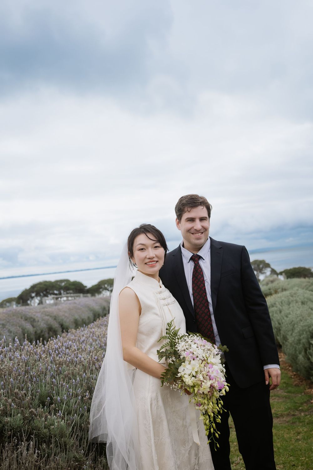 A newlywed couple stands together in a lavender field with a scenic coastal backdrop.