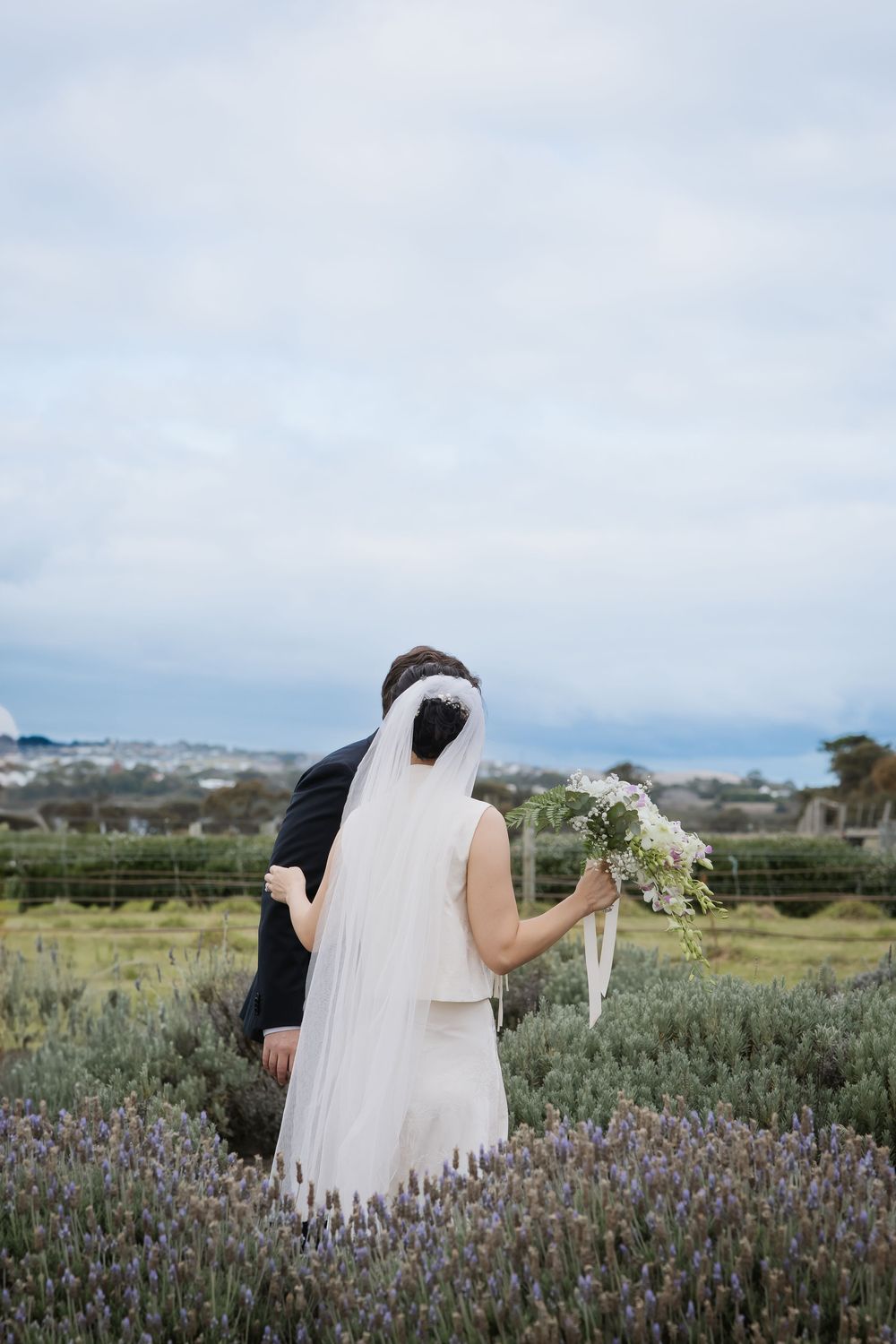 A wedding couple walks through a field of blooming lavender flowers under cloudy skies.