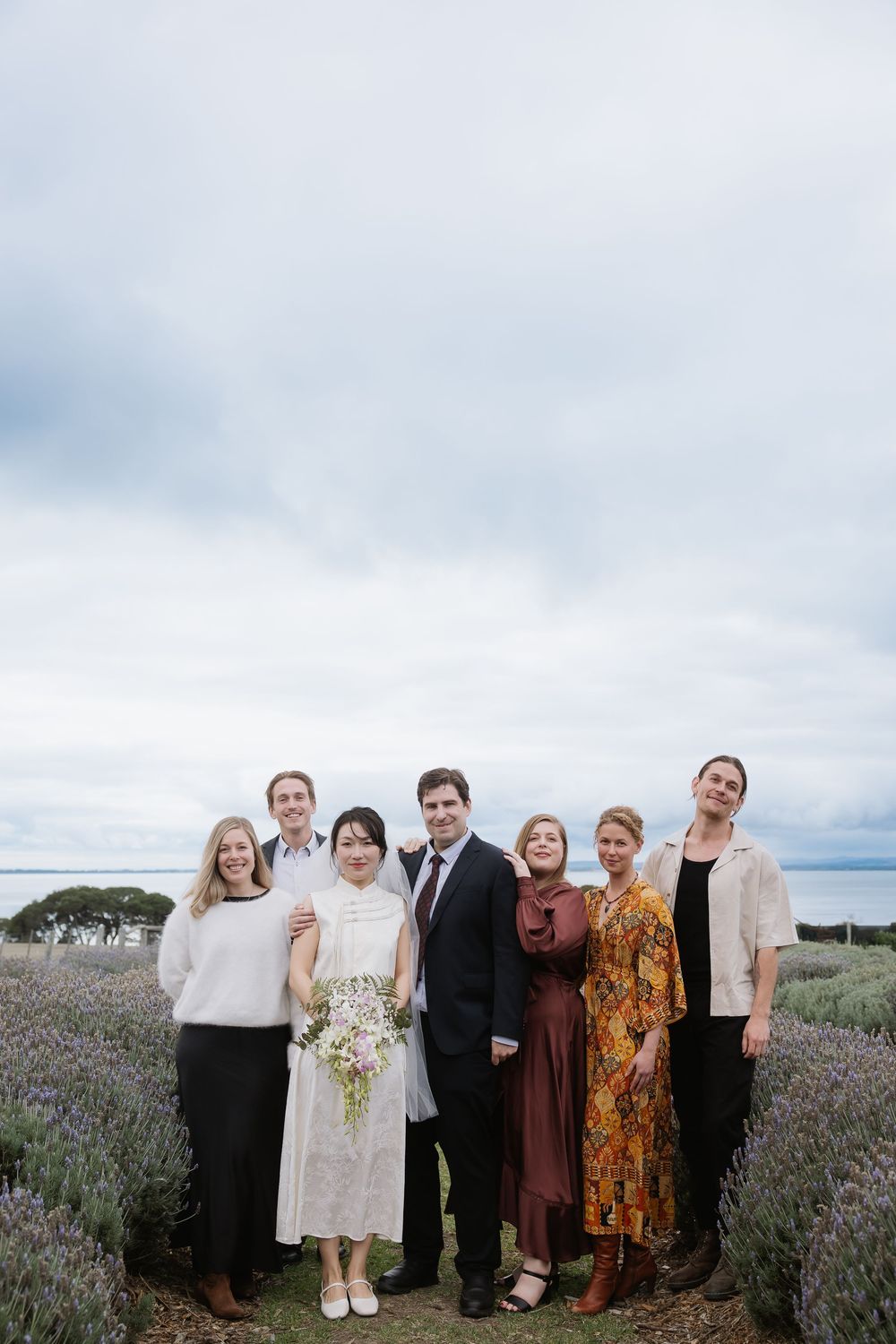 A small group gathers for a photo in a flowering lavender field under cloudy skies.