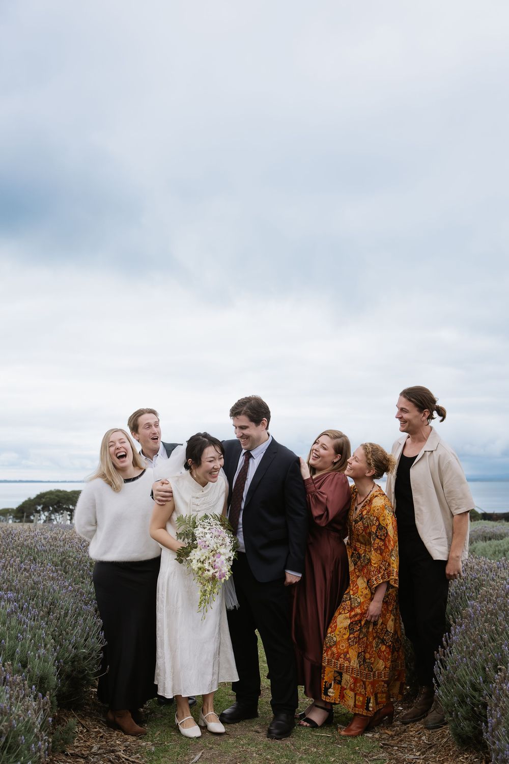 A wedding party shares a joyful moment together in a lavender field under cloudy skies.