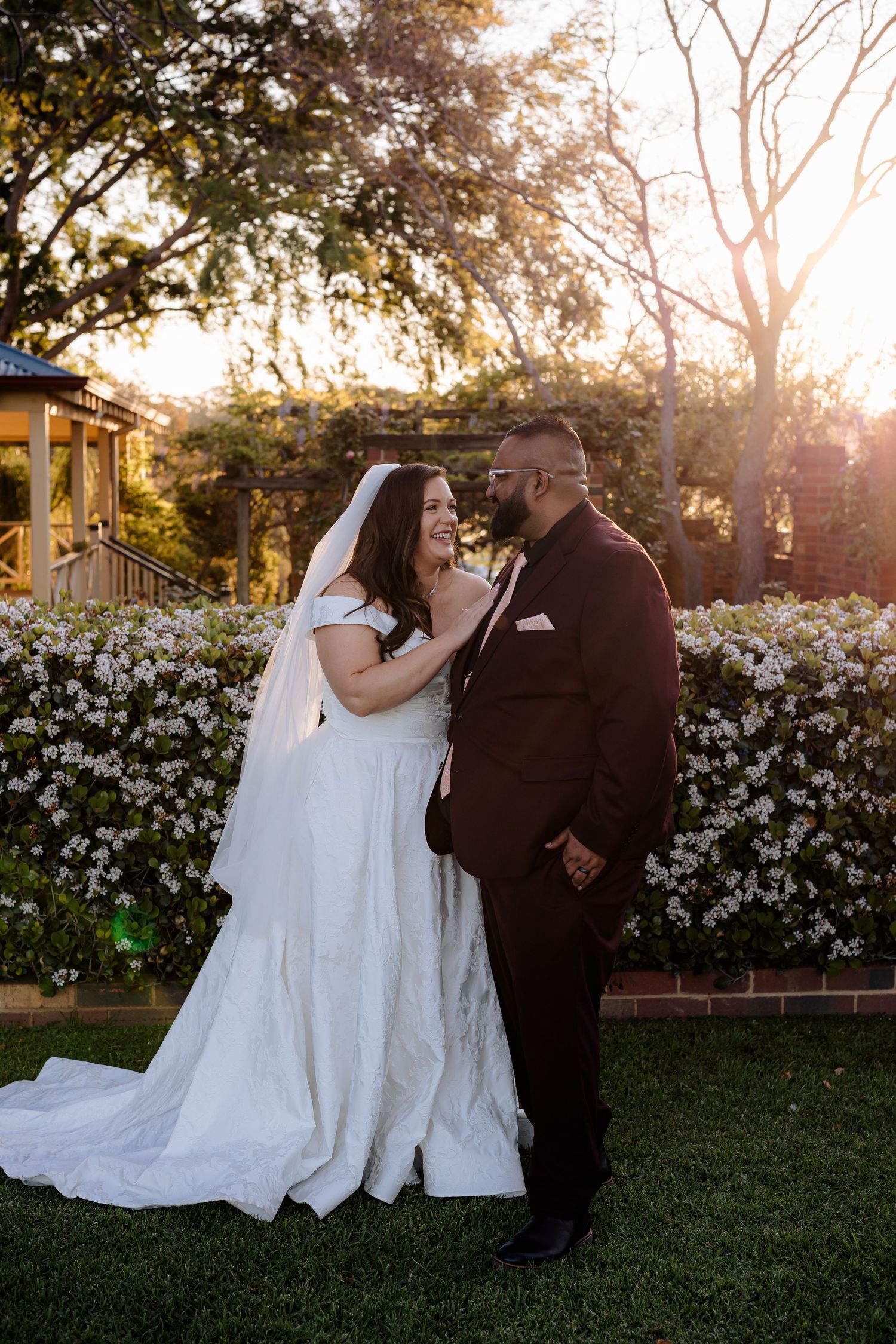 Newlyweds share intimate moments during golden hour by a blooming white flower garden at their romantic outdoor wedding.