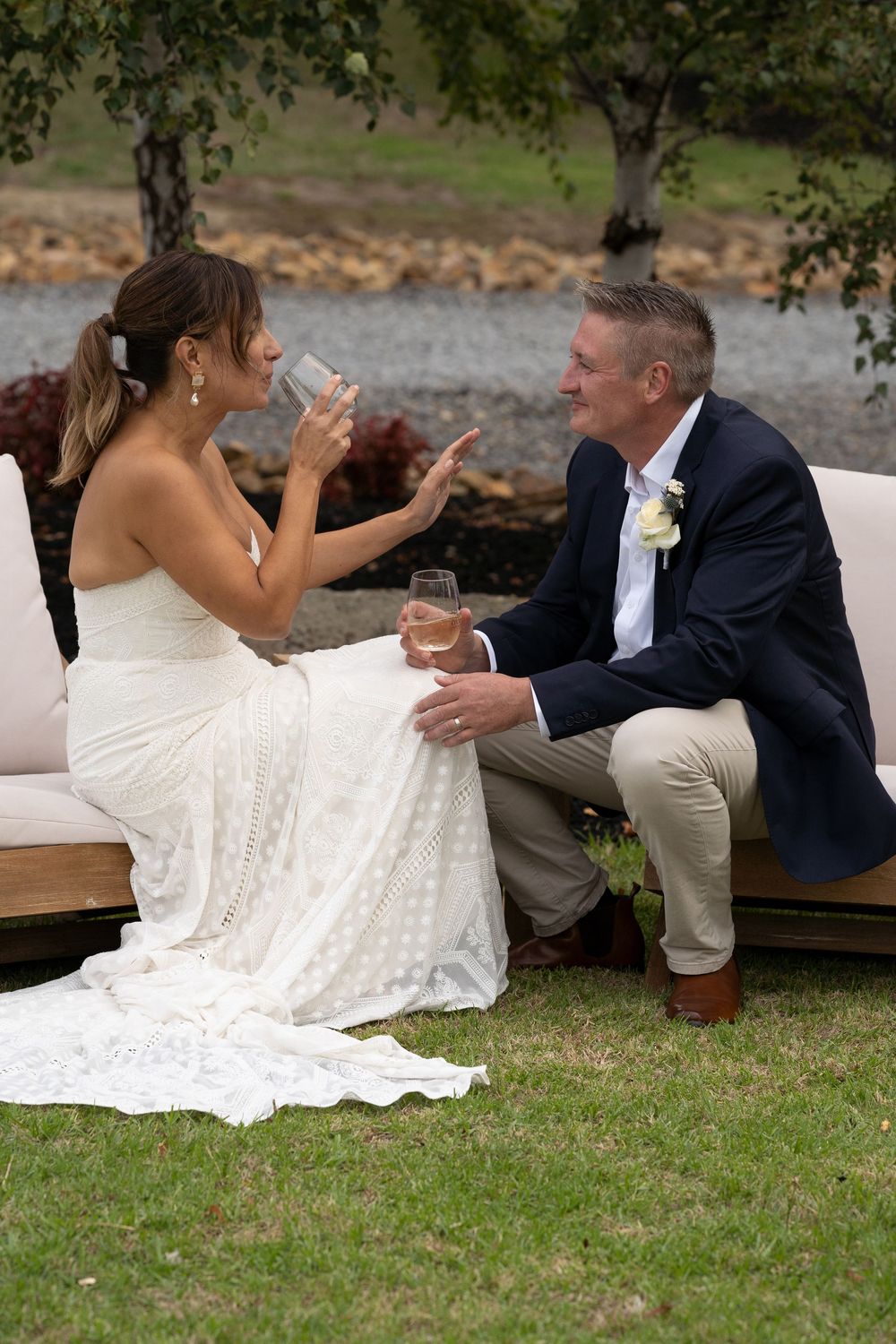 A couple shares a drink on a bench outdoors during a wedding celebration, with a beautiful white lace gown on display.