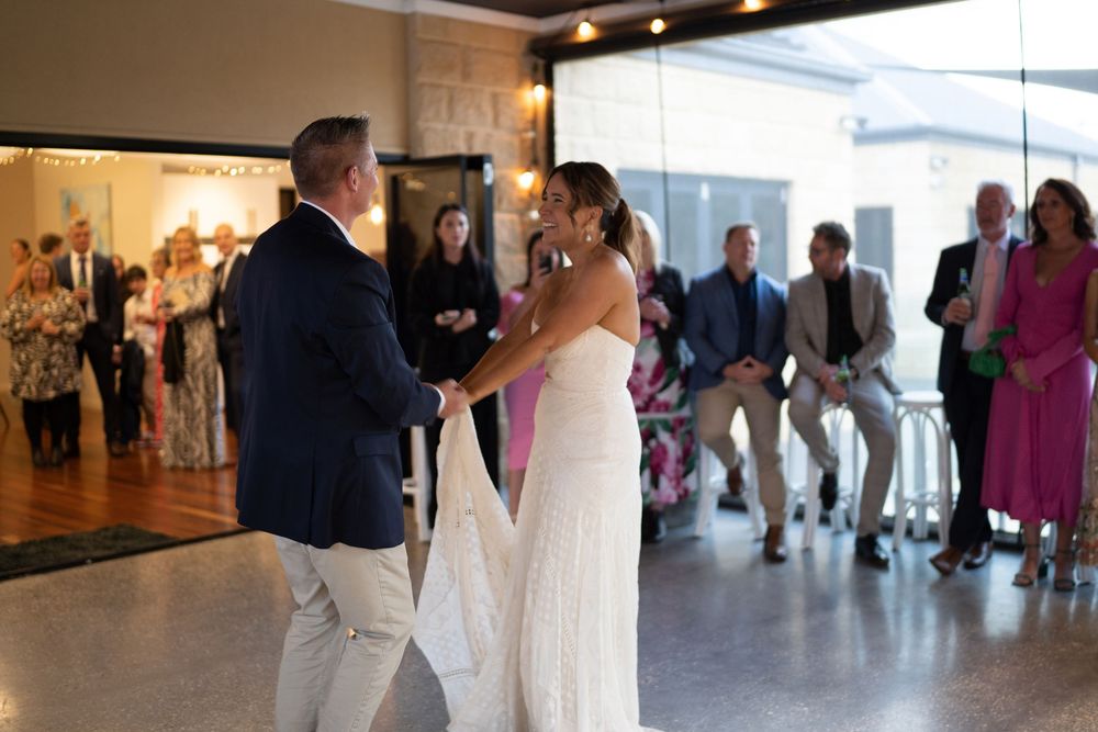 Wedding guests mingle in a modern reception venue with high ceilings and natural light filtering through windows.