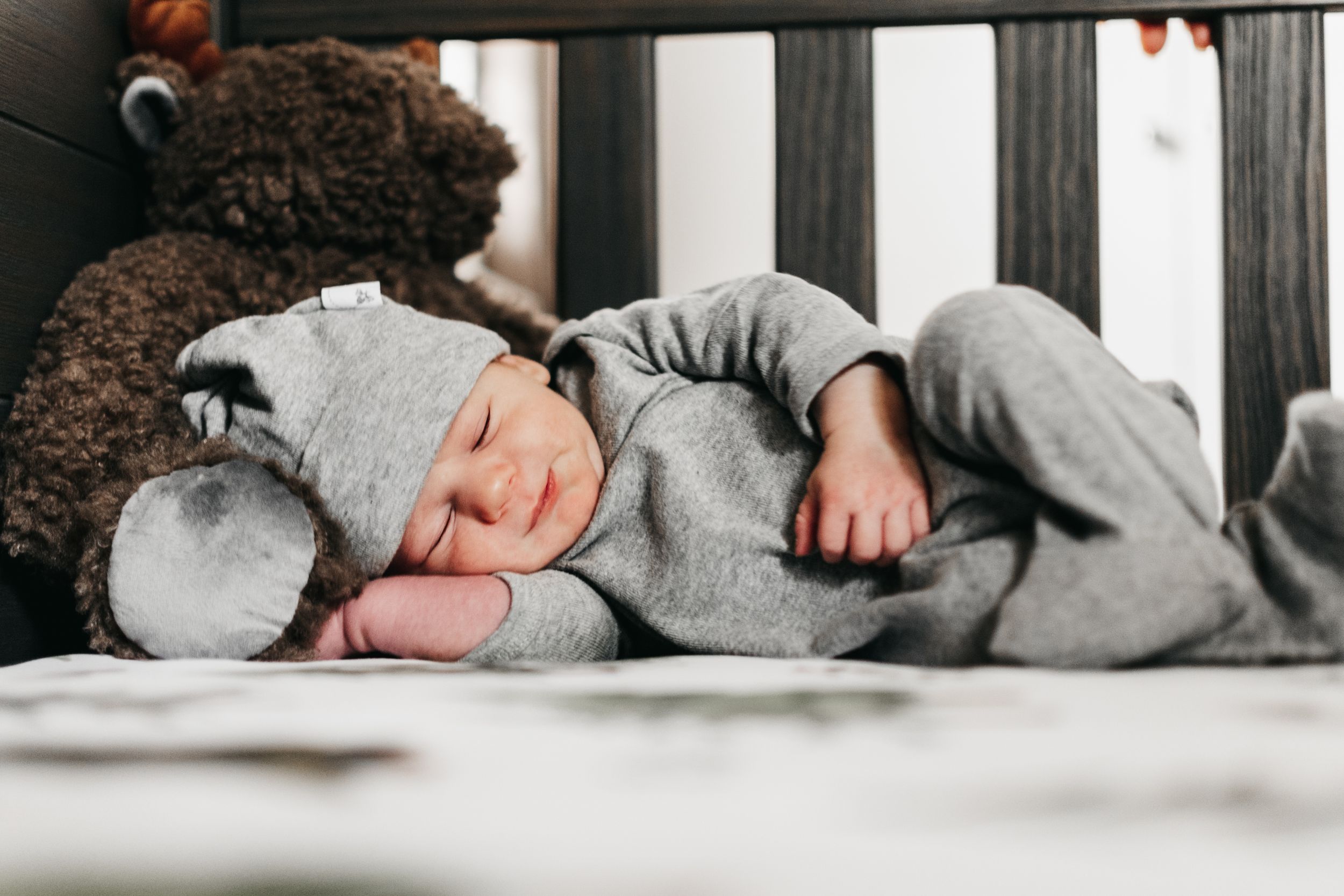 Baby sleeping peacefully in grey outfit next to teddy bear in crib.  Indoor Newborn Session Arlington Texas