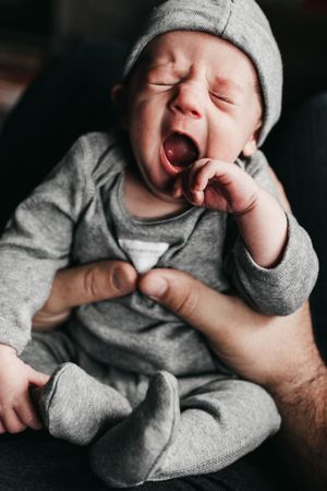 A yawning baby wearing a gray onesie and beanie stretches while being held. Indoor Newborn Session Arlington Texas