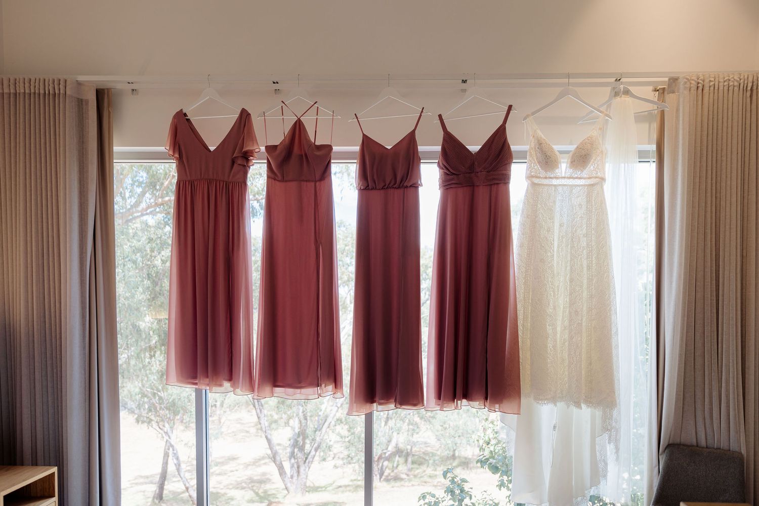 Bridesmaid dresses in shades of burgundy and one white dress hang on a curtain rod in front of a window.