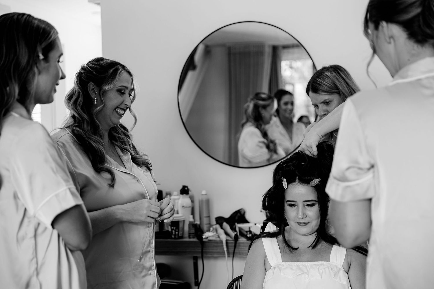 A black and white photo of bridal preparations in a dressing room with a large mirror, hair styling equipment and people getting ready.