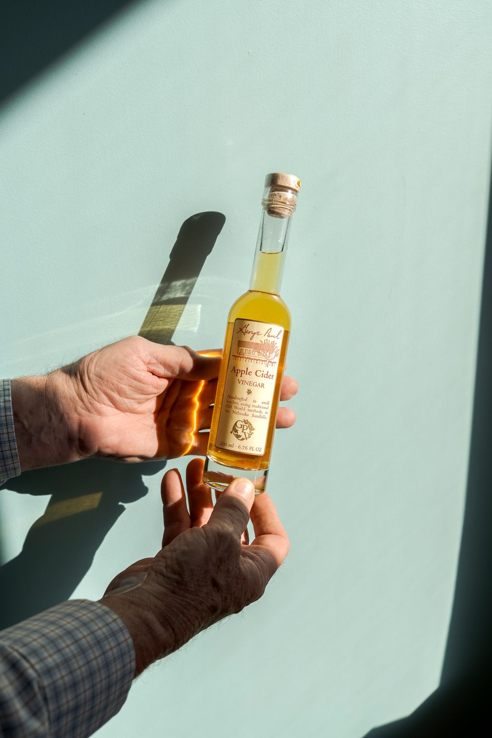 Hands holding amber-colored vinegar bottles against light blue background with dramatic shadows.