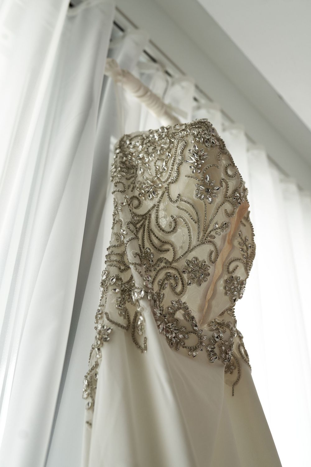Close-up of intricate beaded detail on white wedding dress hanging by window.