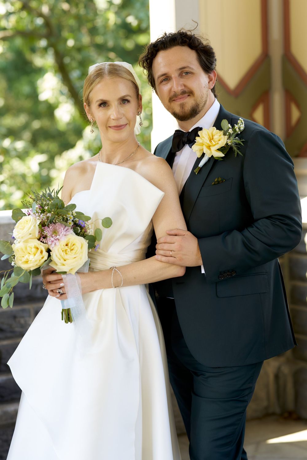 Newlywed couple in formal wedding attire standing close together with yellow floral bouquet.