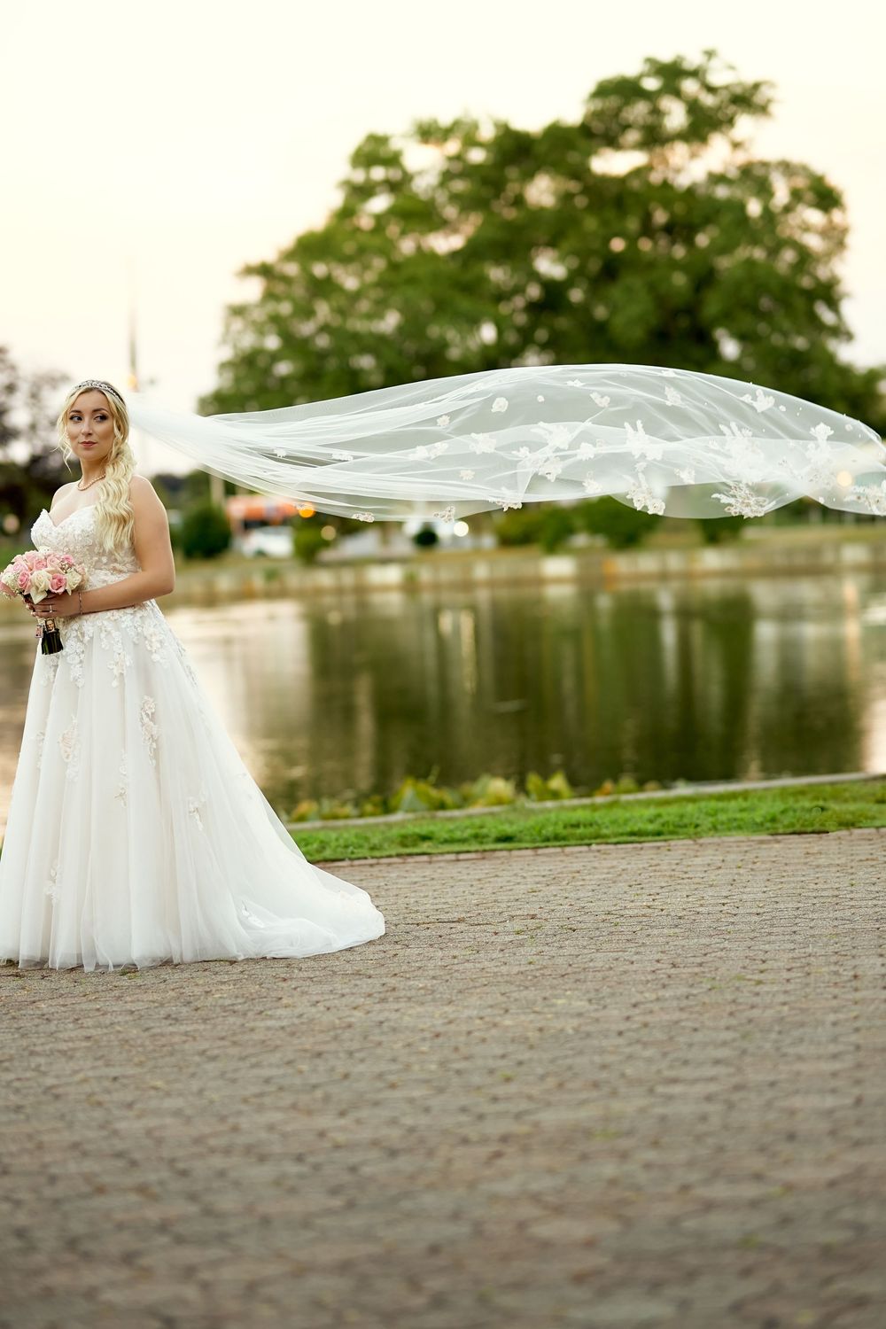 Bride in flowing white dress with veil flowing in wind by lakeside on wedding day.