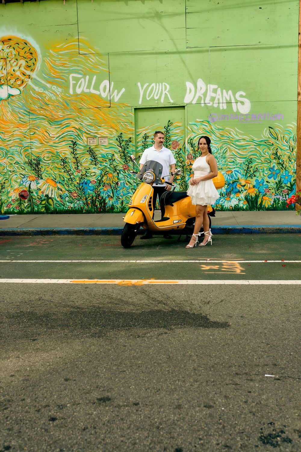 A couple poses with a yellow scooter in front of a colorful mural that says 'Follow Your Dreams'.