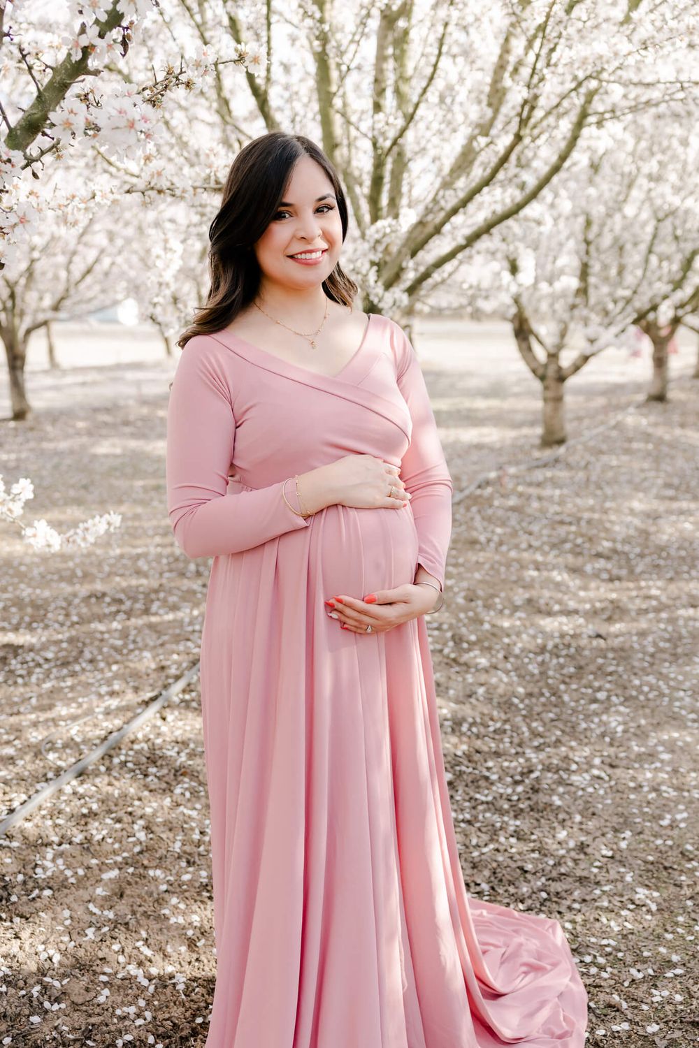 Woman in pink dress poses for maternity shoot among blooming cherry blossom trees, Livermore, CA