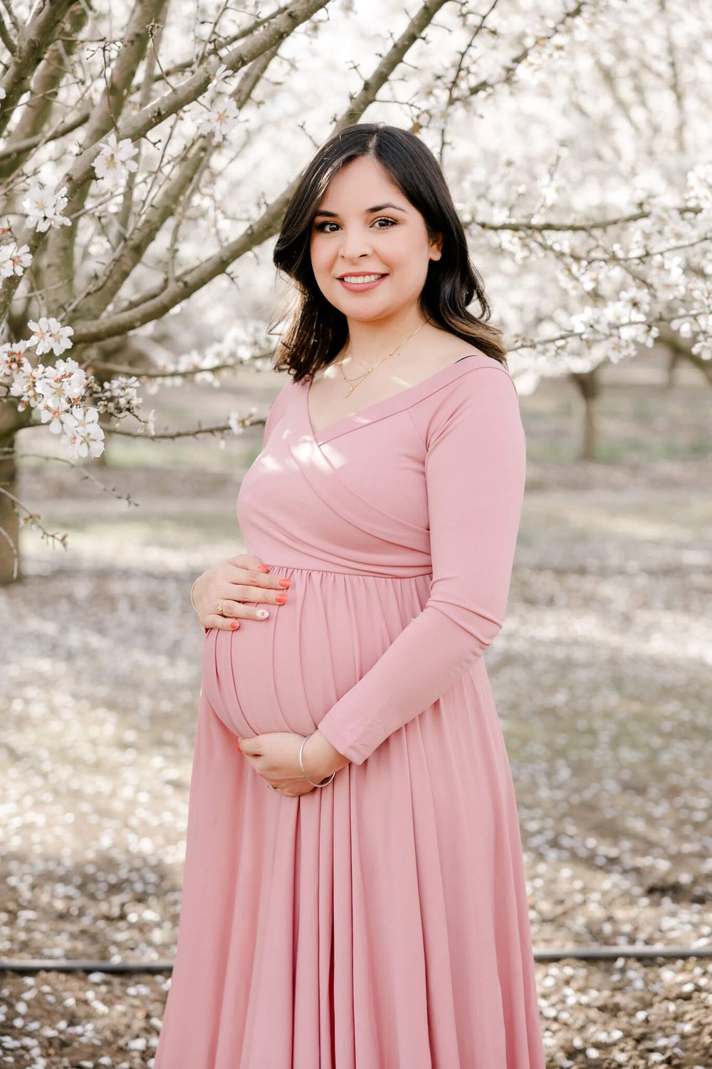 Woman in pink dress poses for maternity shoot among blooming cherry blossom trees, Livermore, CA