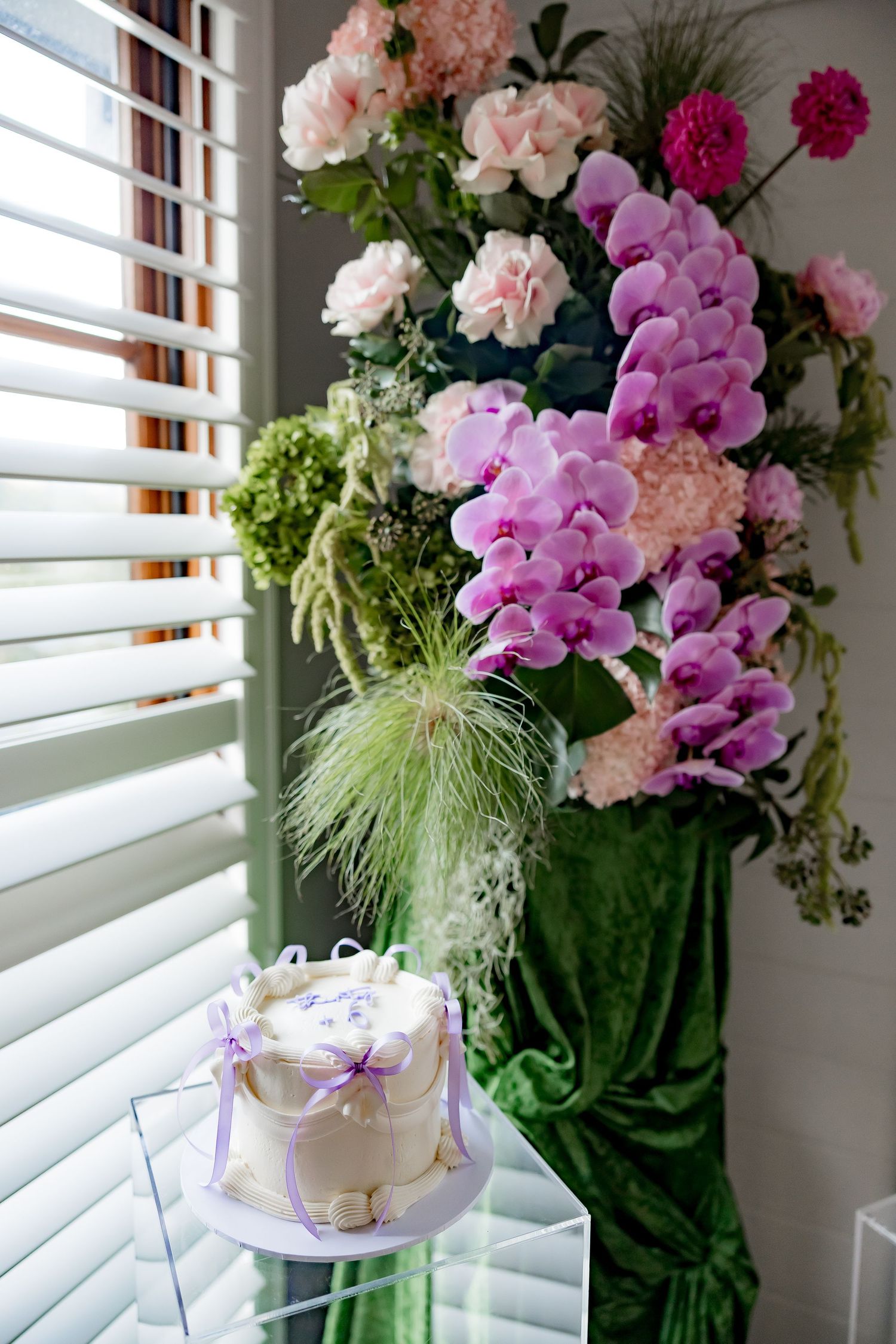 Elegant floral arrangement with pink carnations, purple orchids and feathery grass displayed near a sunlit window.