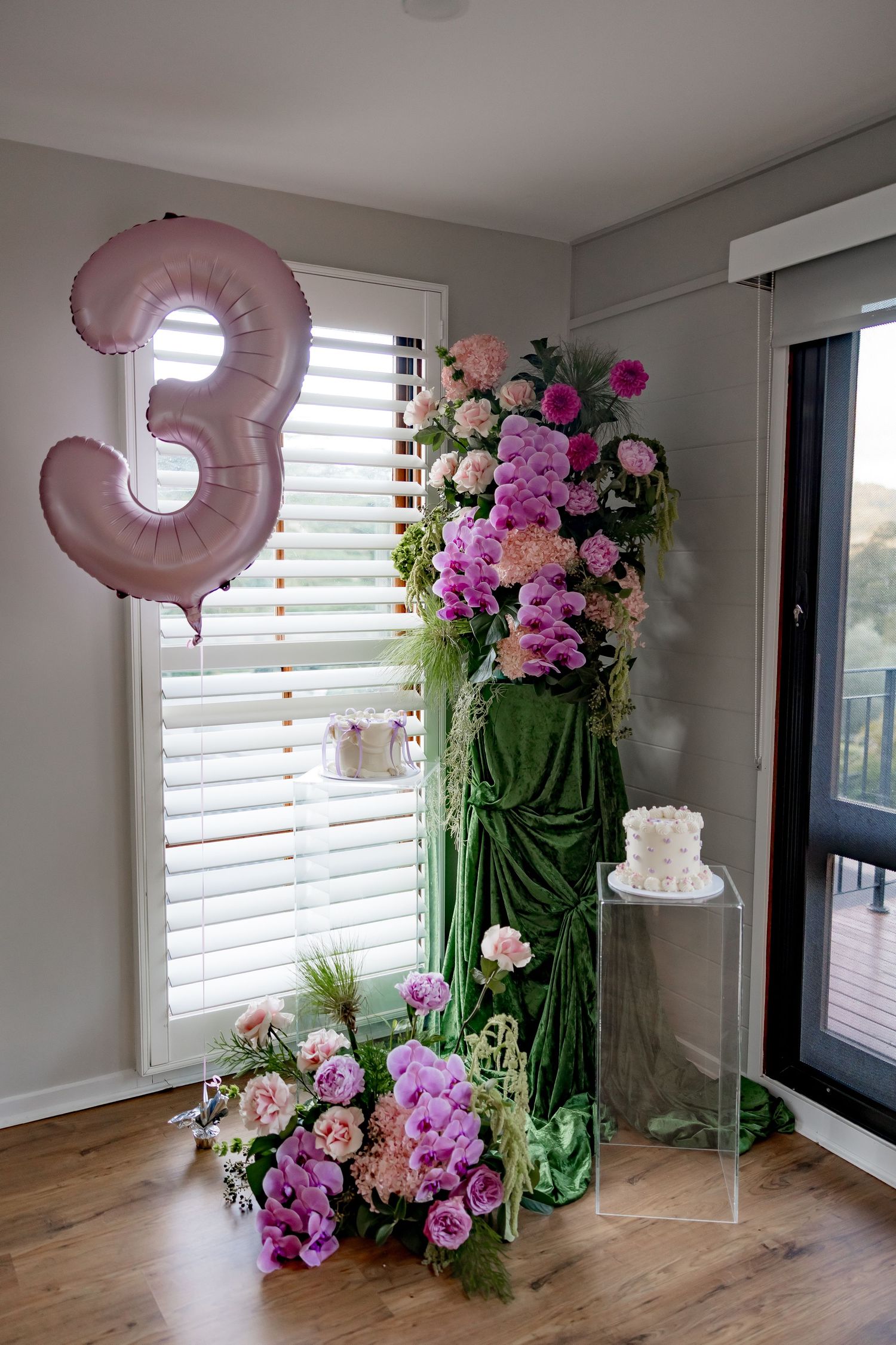 A pink number 3 balloon with elegant floral arrangements in pink and purple tones next to a small white cake on a pedestal.