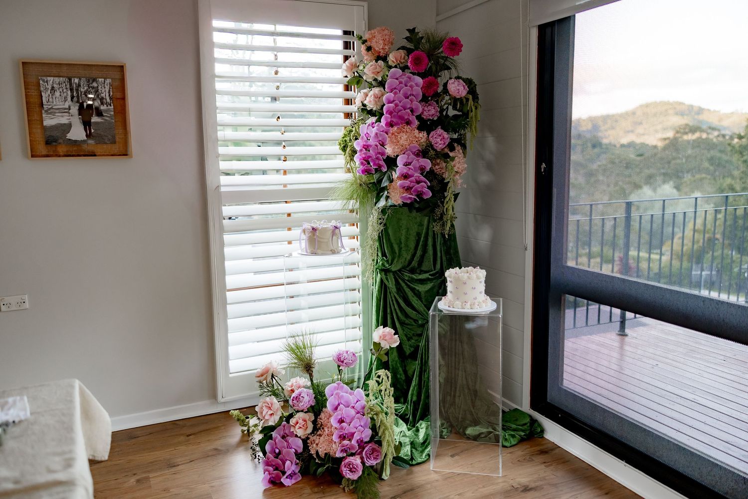 A tall green bamboo vase with pink and purple orchids stands in a corner room with mountain views through the window.