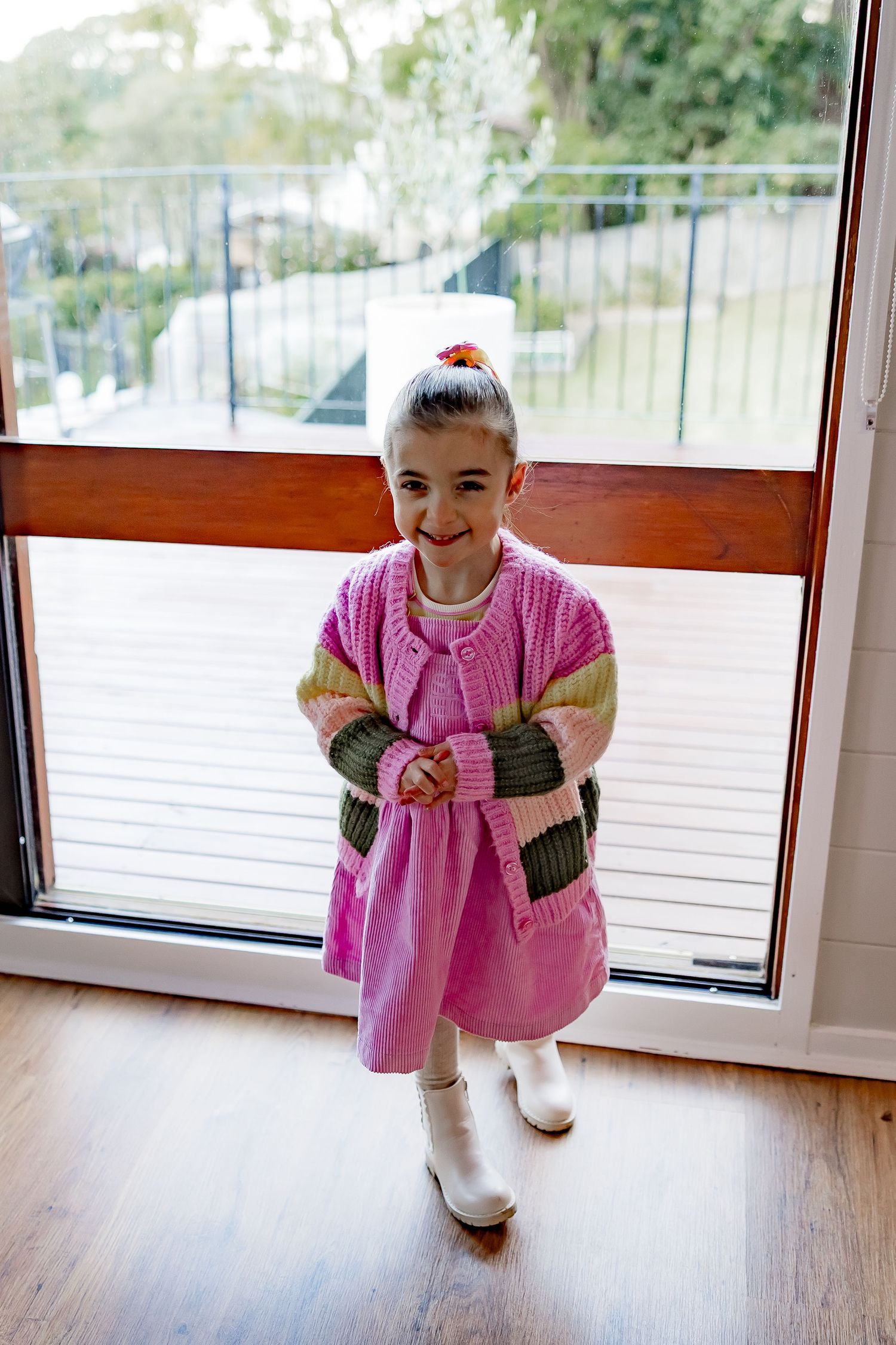 A small child wearing a bright pink dress and white boots stands indoors near a glass door.