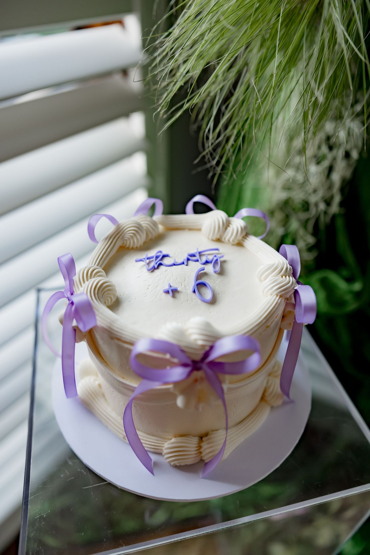 Elegant white round cake decorated with gold trim and purple ribbons displayed near a window with greenery visible.