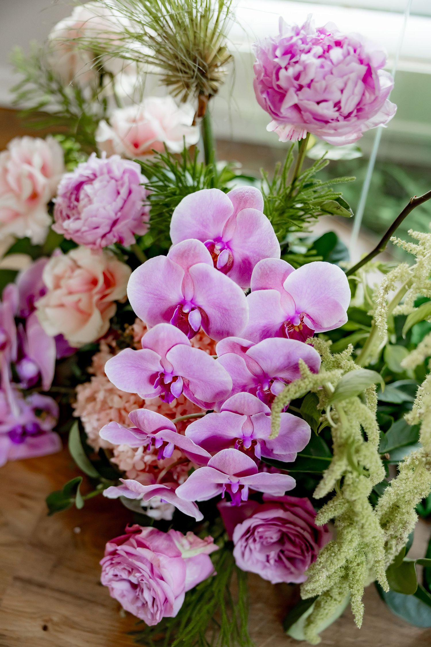 Pink orchids and roses arranged with greenery on a wooden surface in a romantic floral display.