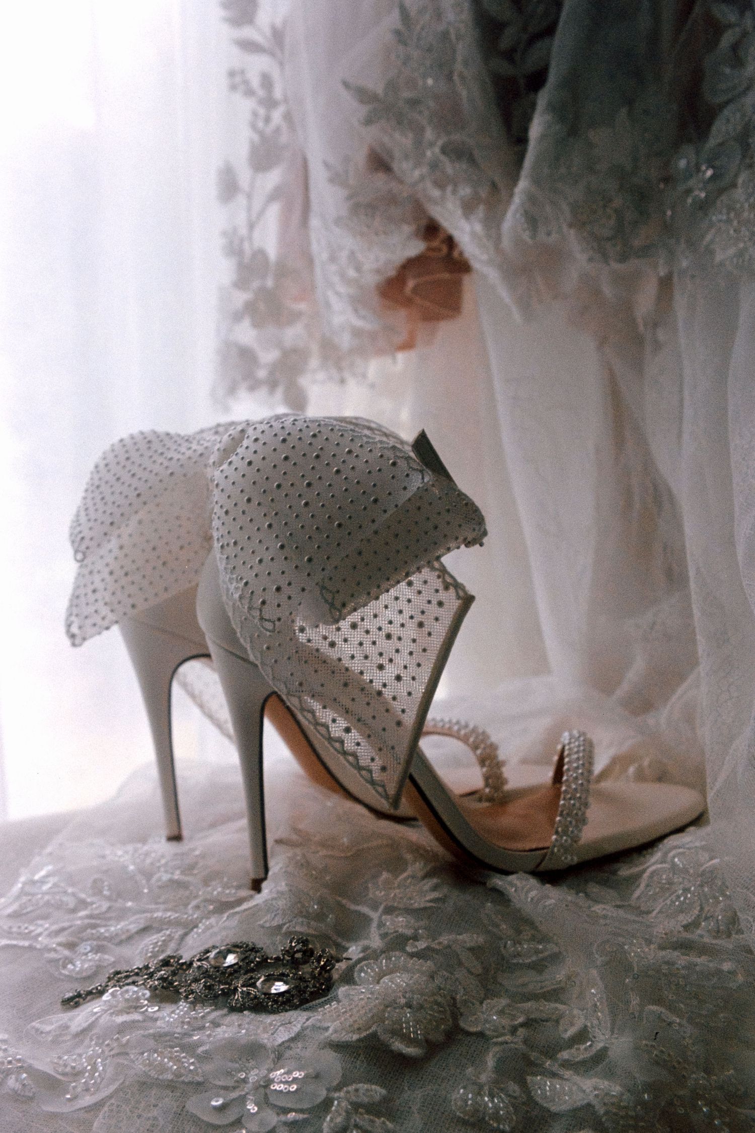 Elegant white bridal shoes with delicate pearl detailing rest on lace fabric in soft ethereal lighting.