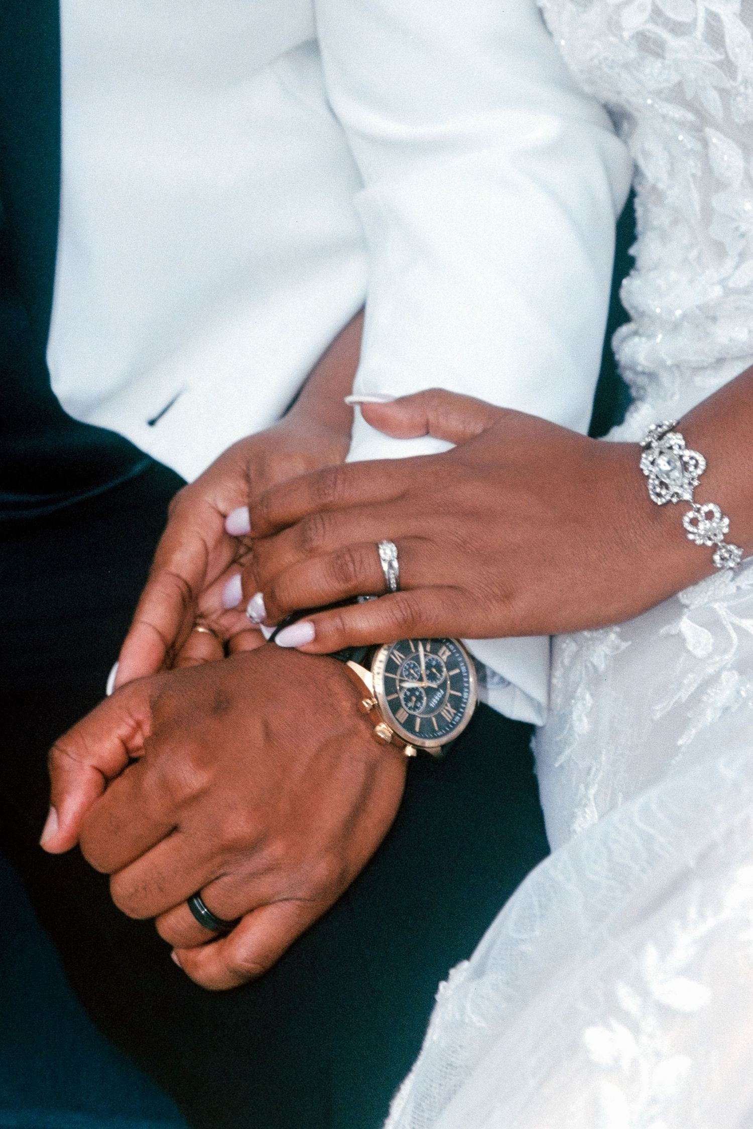 Close-up of hands showing wedding rings, with white lace dress fabric and elegant silver bracelet details.