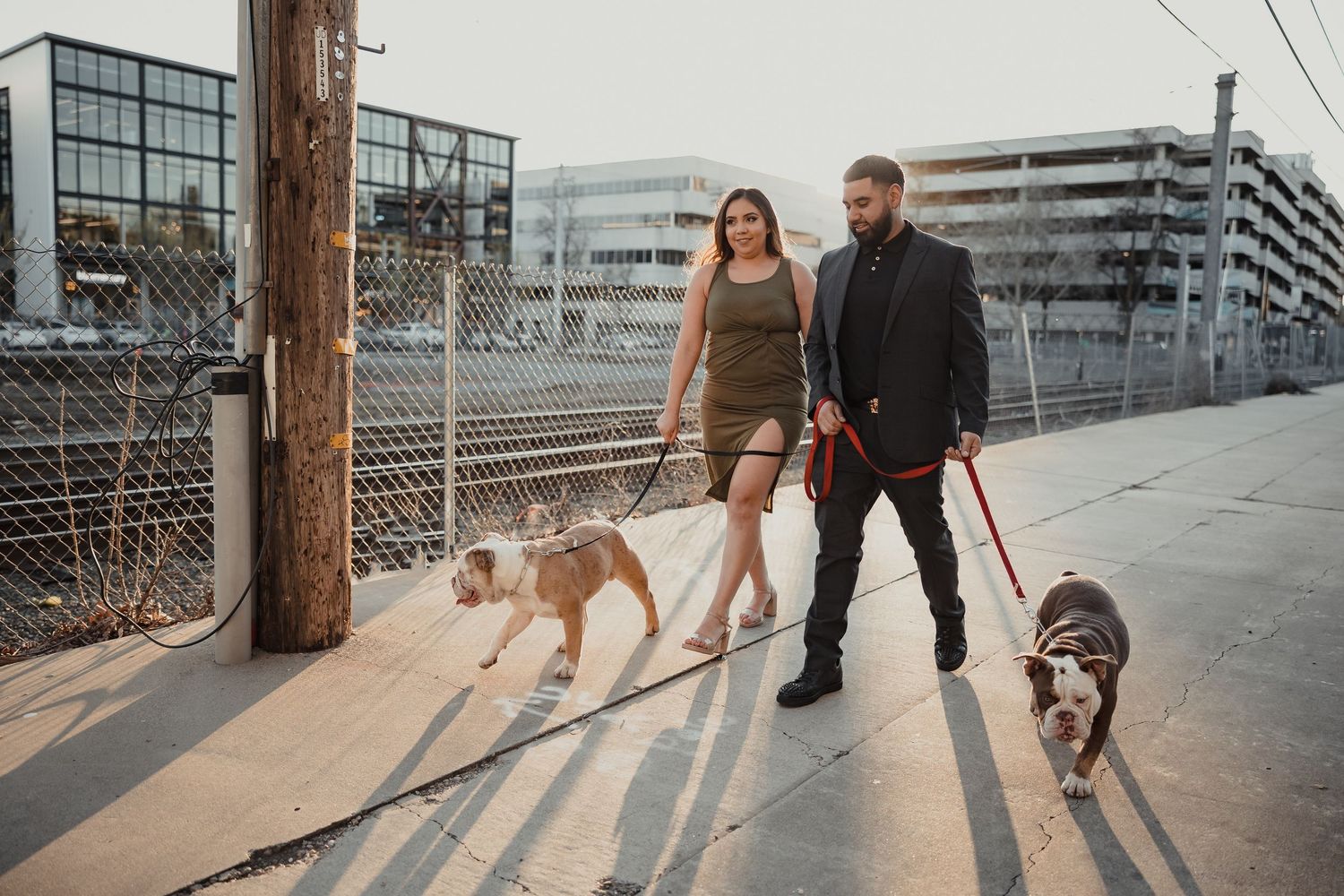 A couple walking their two dogs together on an urban sidewalk at sunset, with commercial buildings in the background.