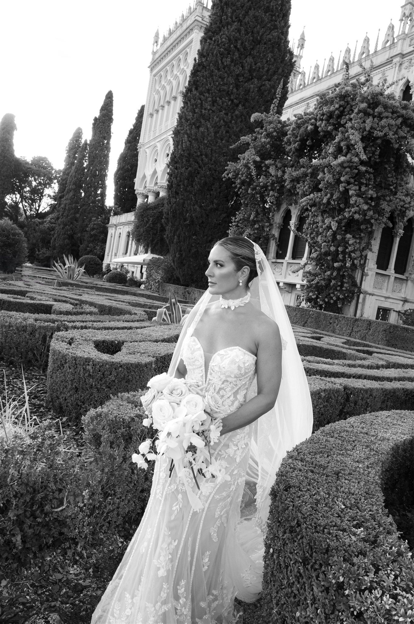 Black and white photo of a bride in a flowing dress posing in a formal garden setting.