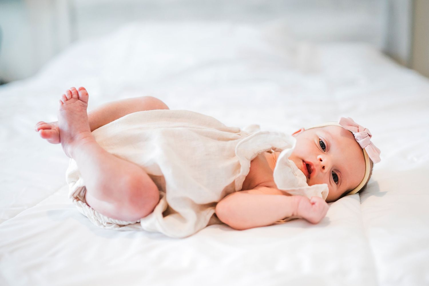 Happy baby in white onesie lying on white bedding while laughing and playing.