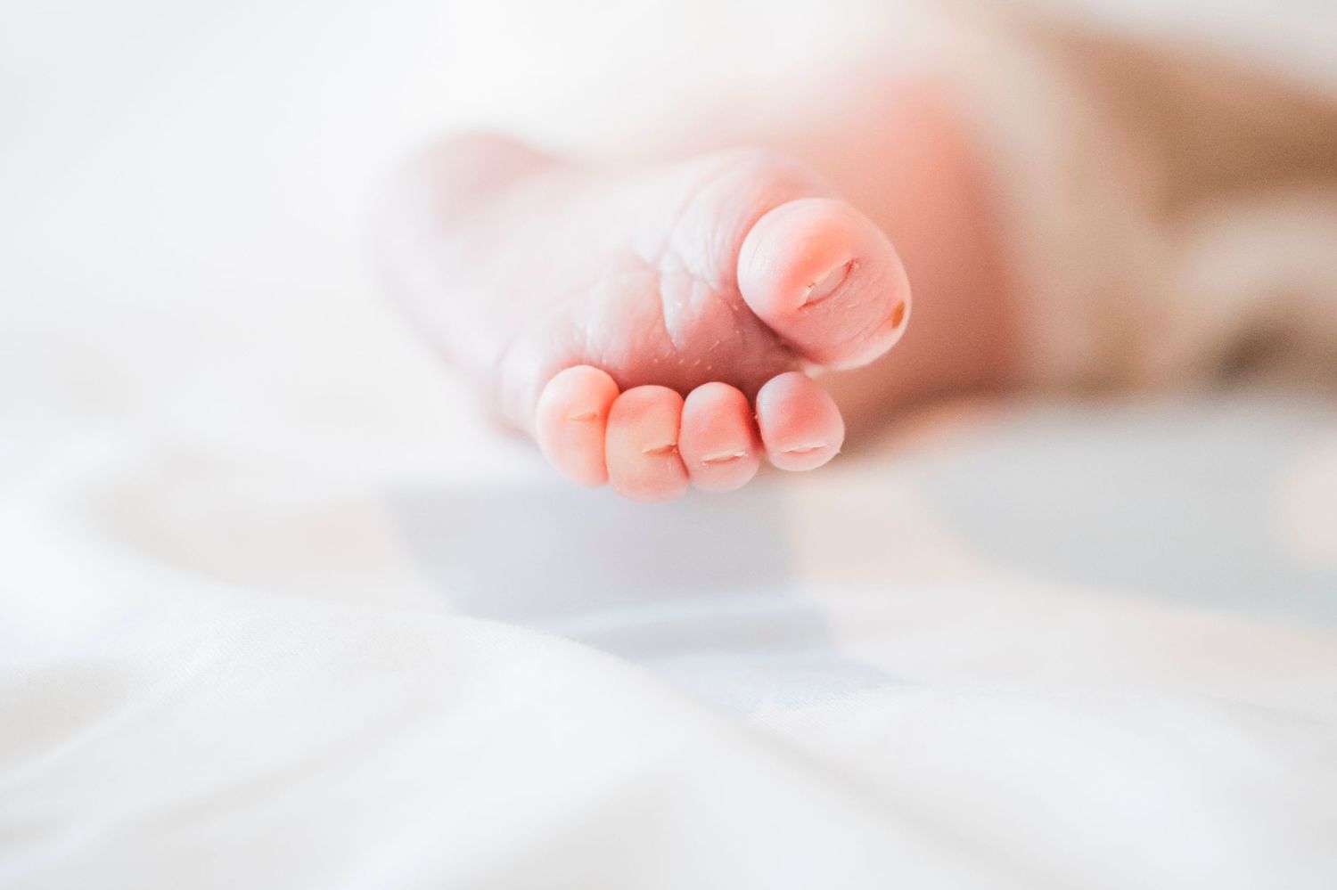 Tiny newborn baby toes resting on soft white sheets in gentle natural light.