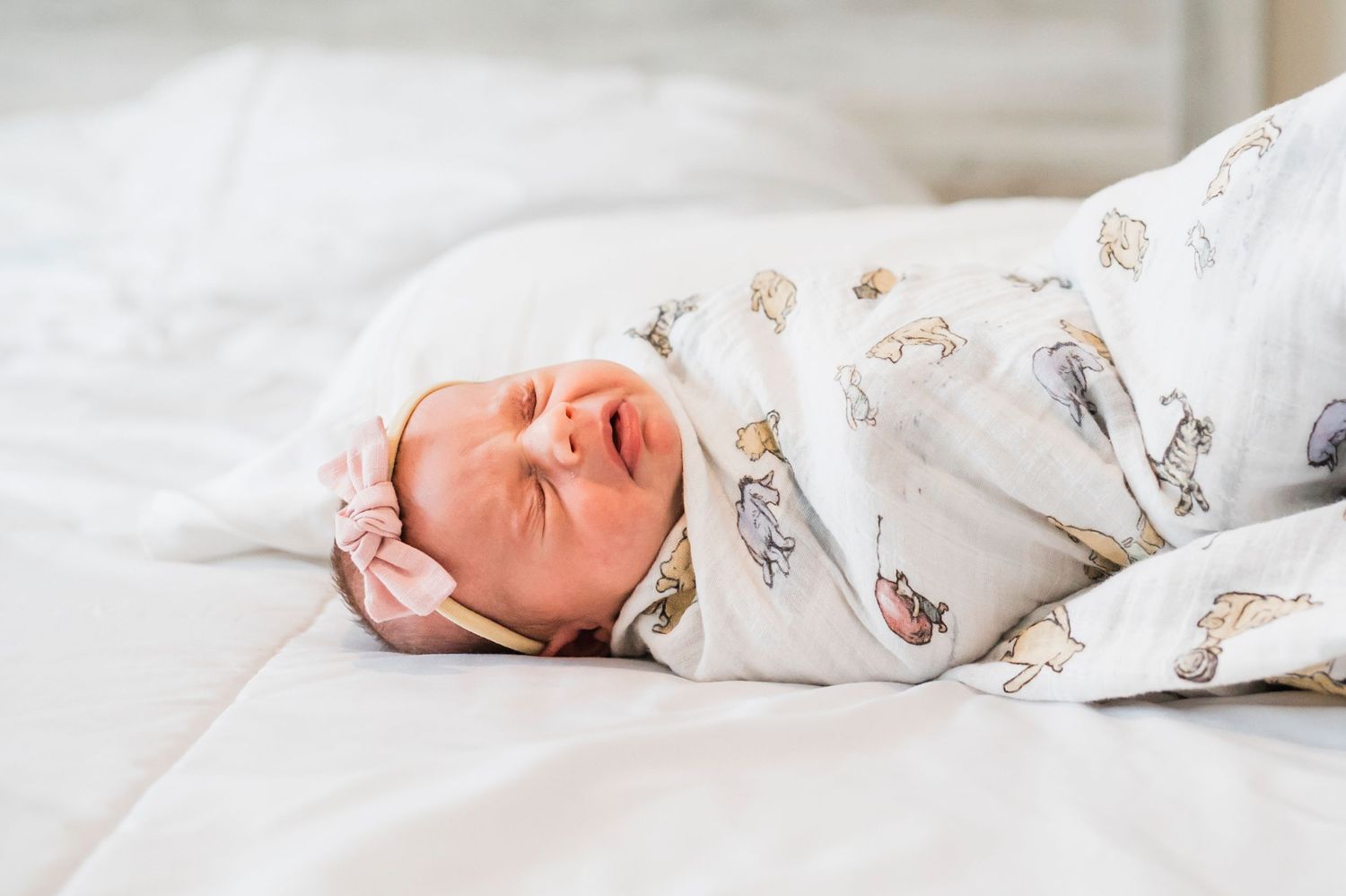 A newborn baby wearing white sleepwear lies on soft white bedding while sleeping peacefully.