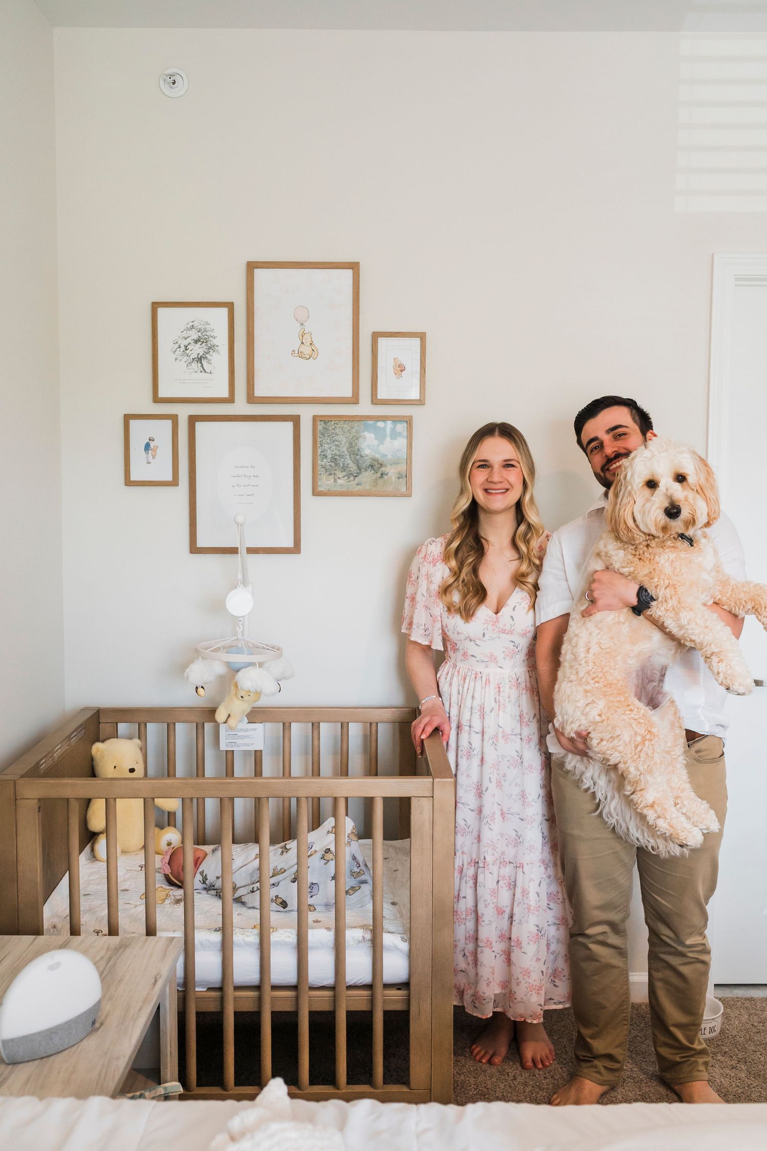 A cozy nursery room with a wooden crib, framed wall art, and a couple standing with their fluffy goldendoodle dog.