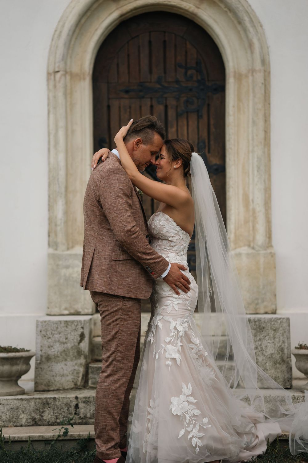 A couple in wedding attire share an intimate moment in front of an arched wooden church door.