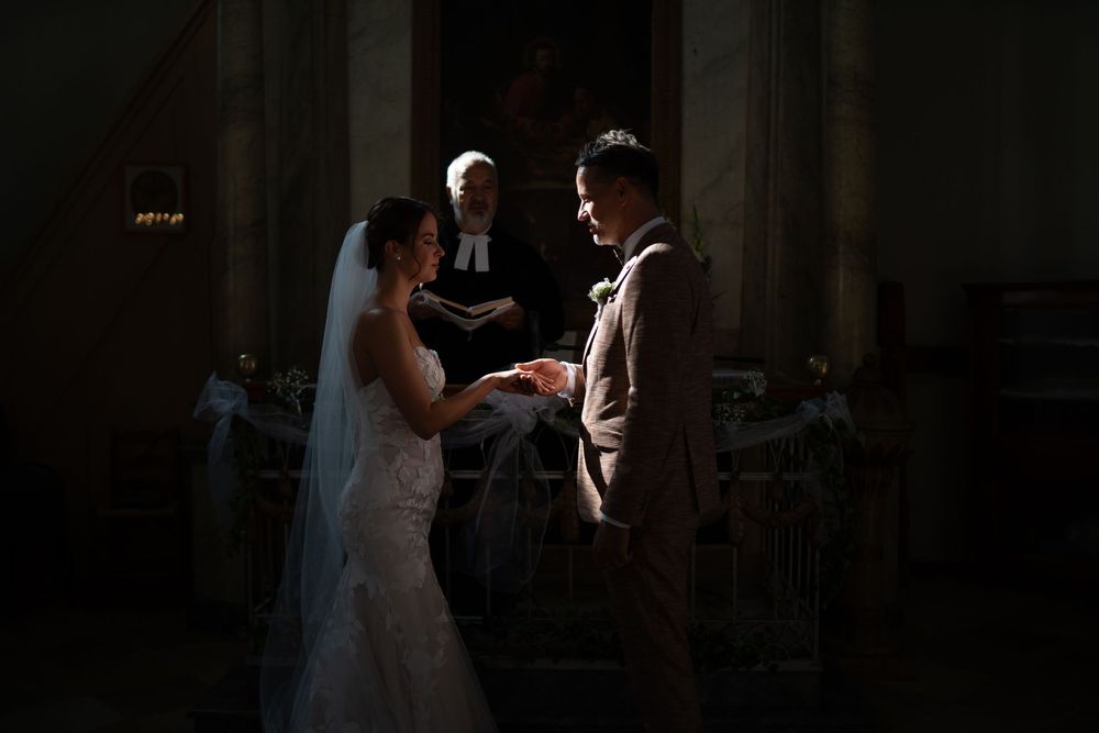 A couple exchanges vows during their wedding ceremony in a dramatic, shadow-lit church interior.