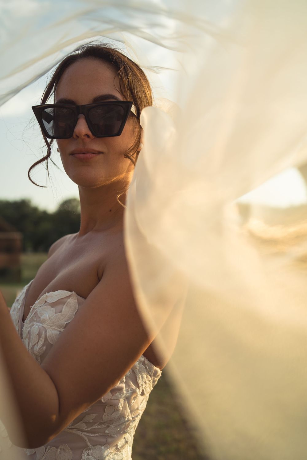 Bride in sunglasses and lace wedding dress poses during golden hour while veil flows in breeze.