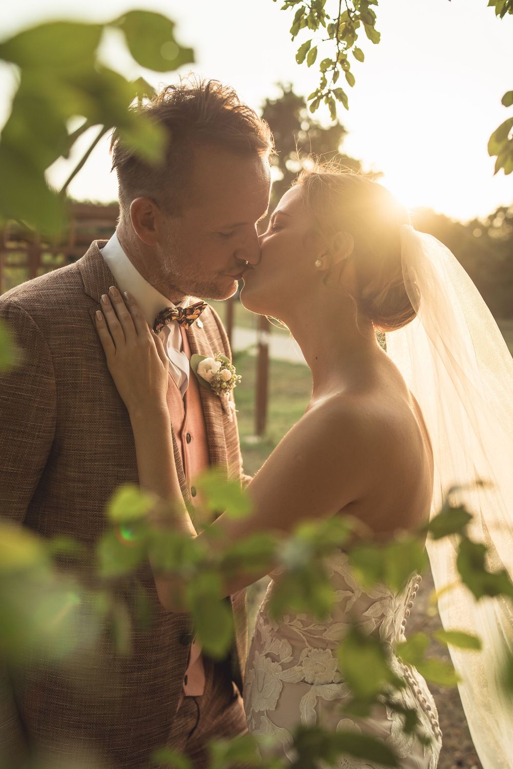 Romantic wedding couple shares an intimate kiss at sunset through green leaves in a natural outdoor setting.