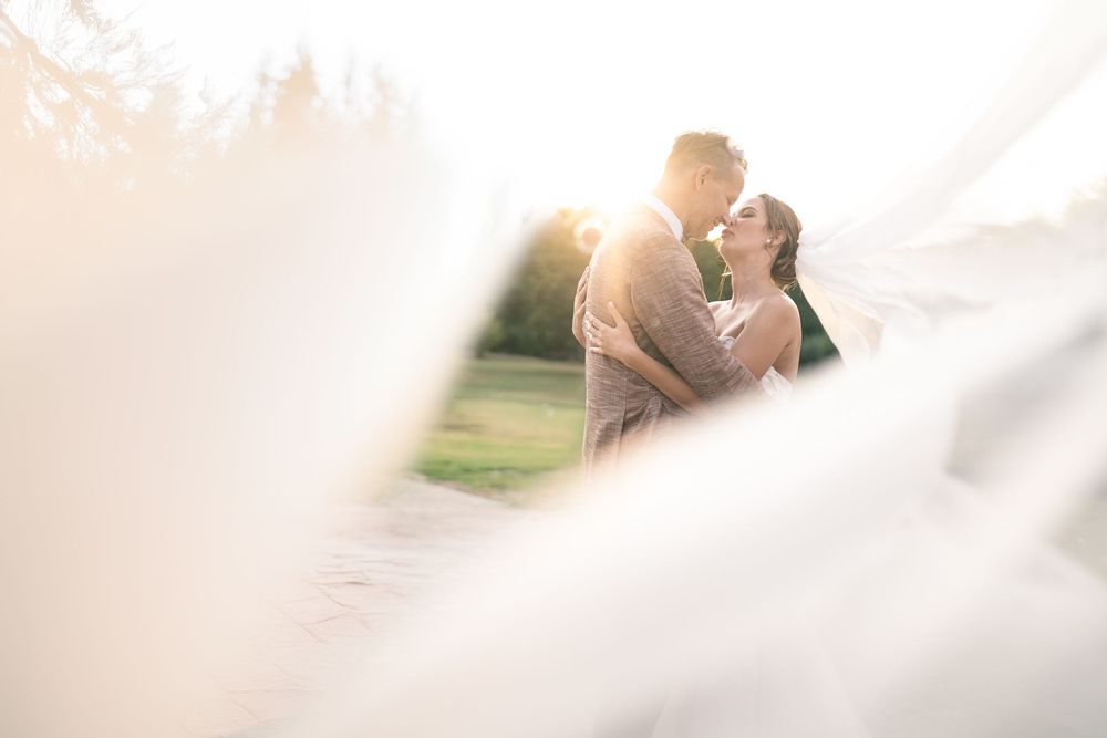 A romantic couple in wedding attire embraces passionately in a sun-filled outdoor setting.
